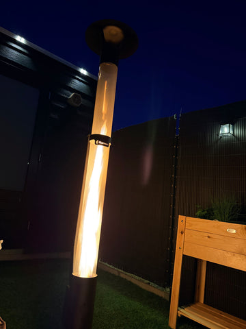 Outdoor pellet heater burning at dusk next to a wooden bench in a green garden